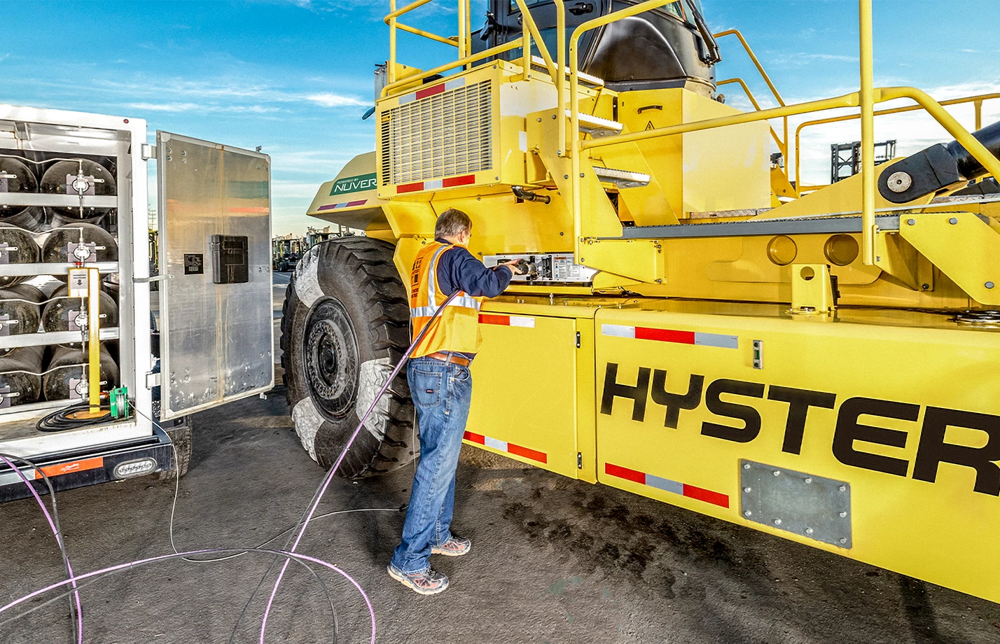 A man refueling a Hyster HFC Reachstacker from a rack of hydrogen tanks at a port facility.  